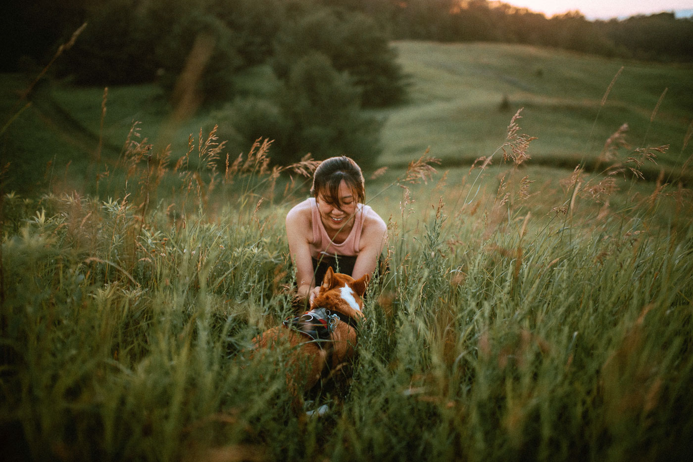 Woman smiling while crouched in meadow petting a corgi that is facing her. Location: McCormack Trail located in Hamilton, Ontario