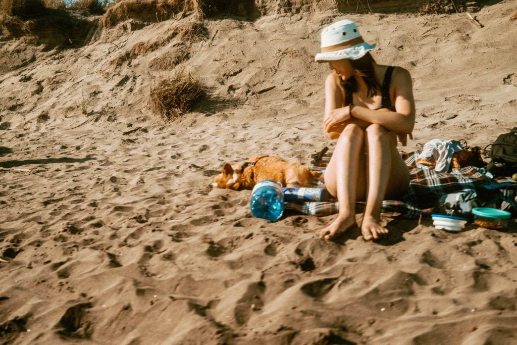 Corgi laying in the sand beside a picnic blanket. A woman in a swimsuit is sitting on the picnic blanket watching the corgi roll in the sand at Long Point Provincial Park's Dog Beach