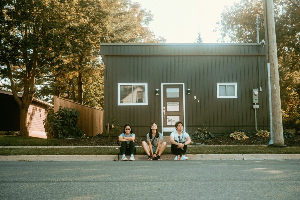 3 friends sitting by the curbside enjoying poutine and burgers in front of a dark grey house. 
