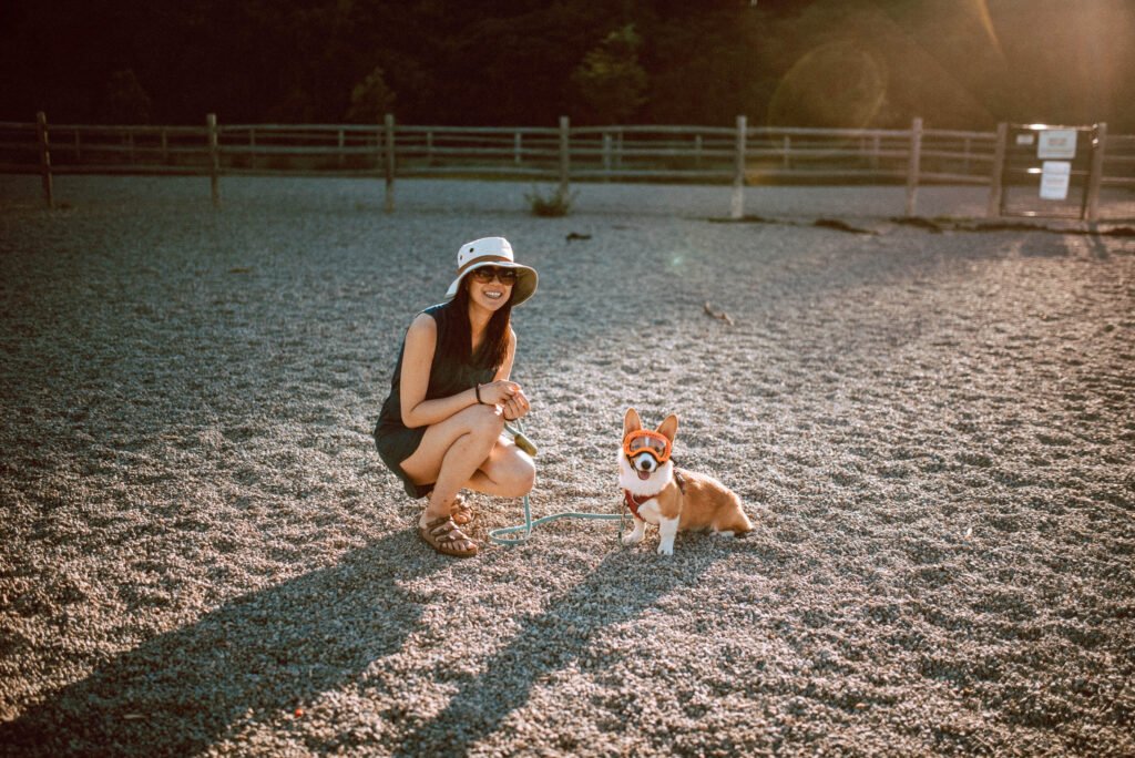 Woman bent down beside red and white corgi wearing a clear Rexspex goggle with orange frames at Raymore Park's Off-Leash Area