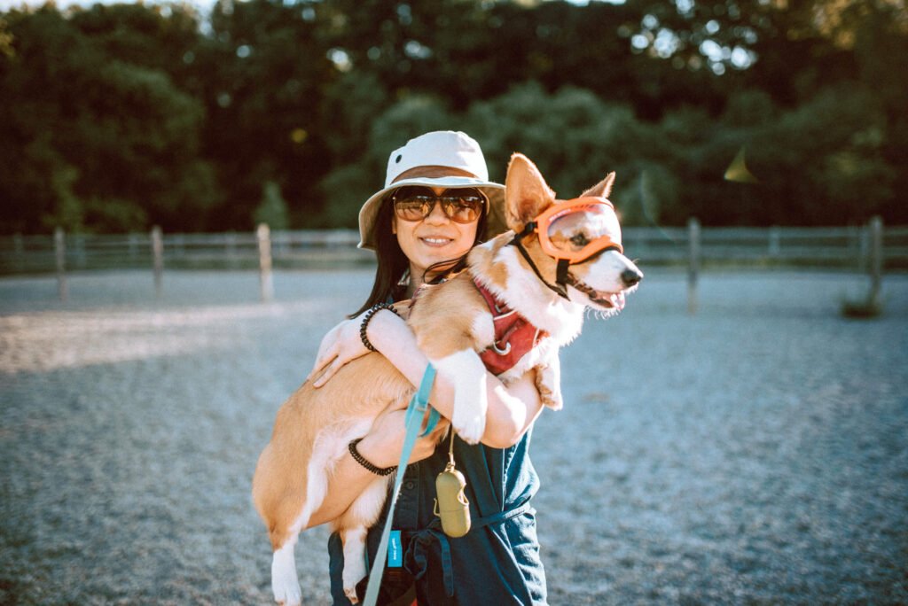 Woman in a soft brimmed hat wearing sunglasses. She is holding a red and white corgi wearing a pink Ruffwear harness and a clear Rexspecs goggle with orange frames