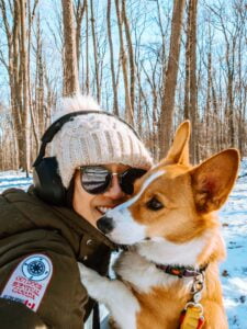 Maria (human) wearing sunglasses, a beanie, and headphones smiling while Limone (red and white Pembroke Welsh Corgi right) offers her cheek for a kiss in the middle of their walk through Baker Wood trails