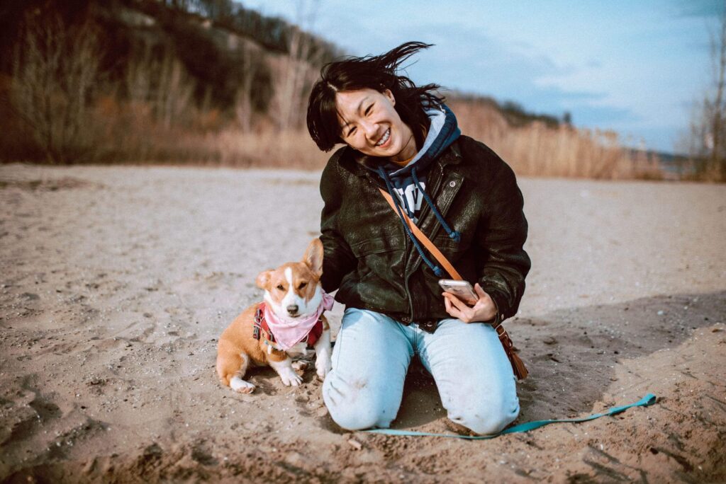 young red and white corgi wearing a pink bandana and harness sitting beside a woman wearing an oversized leather jacket kneeling on the sandy beach at Bluffers Park in March