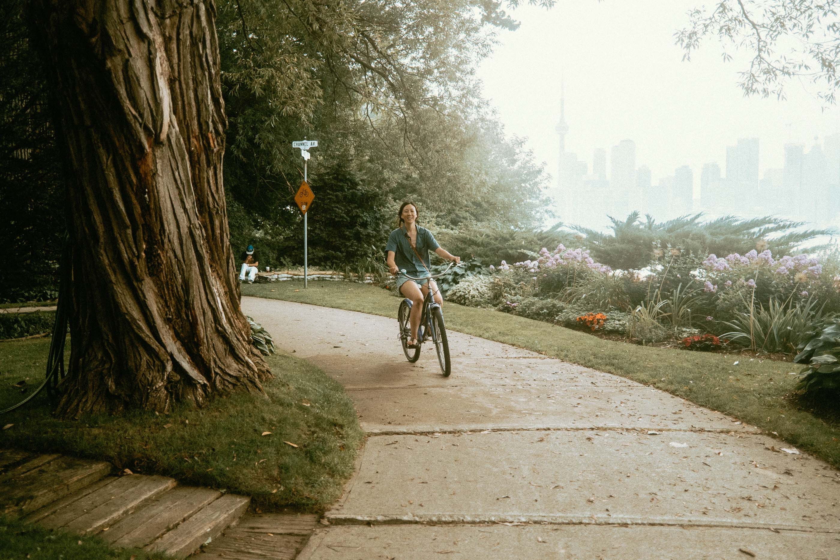 Maria is on a bike rounding a corner of a parkette in Toronto Centre Island - Eastern Channel Parkette. In the distant horizon, you can se the Toronto CN Tower and the city skyline in through the haze.