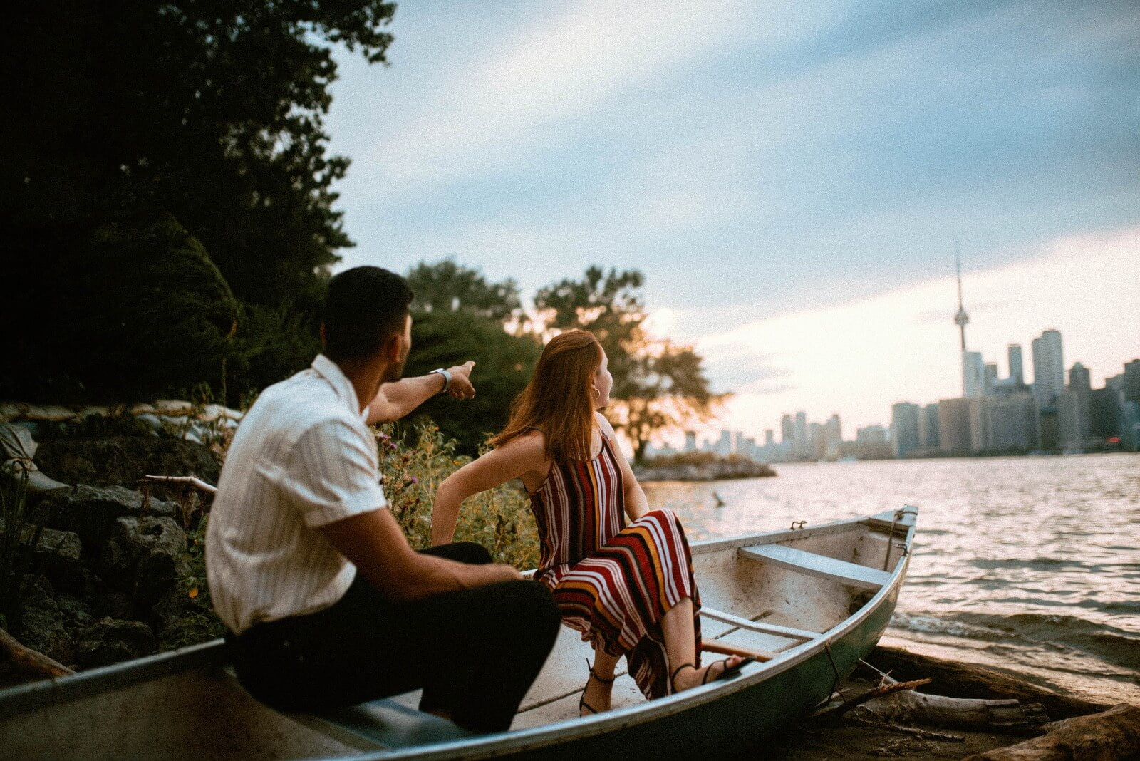 Man and Woman sitting on a small boat at one of the beaches at Toronto Centre Island. The man is pointing to the horizon, where the Toronto city skyline appears in the horizon. Image is taken close to sunset.